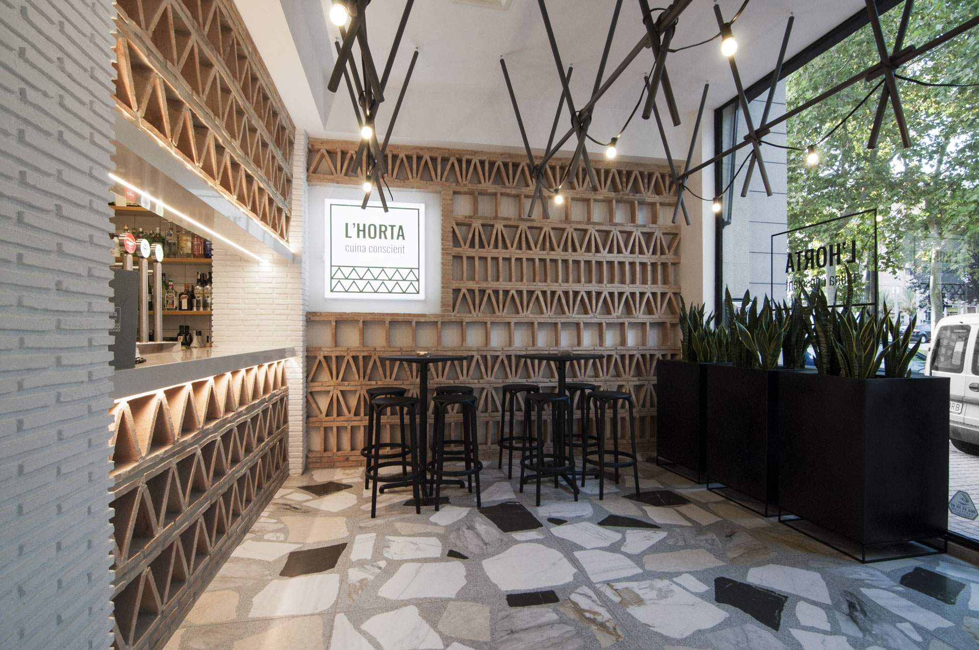 Diseño zona entrada restaurante realizada con ladrillos macizos con colocación según patrones geométricos. Suelo porcelánico de formato 120x120 simulando terrazo. Mobiliario e iluminación suspendida en tonos oscuros. Diseño moderno y elegante.