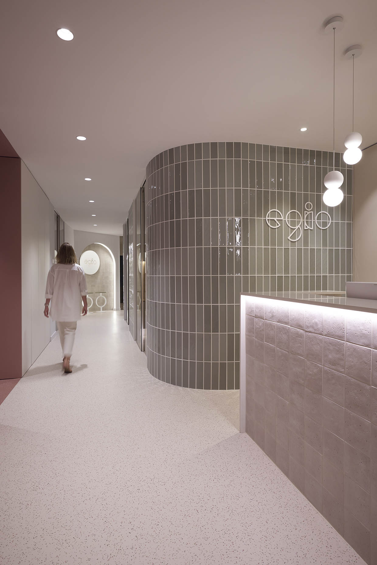 Zona entrada clínica combina materiales y texturas sofisticadas. Destaca una pared curva revestida con azulejos verticales en tono verde grisáceo brillante, contrastando con superficies lisas en tonos neutros. Las puertas de vidrio aportan transparencia y luminosidad, mientras que la iluminación indirecta y los detalles decorativos, como el logo retroiluminado y los elementos circulares en el fondo, refuerzan la identidad visual del espacio. El conjunto transmite una sensación de serenidad y exclusividad, ideal para un entorno de bienestar y salud.