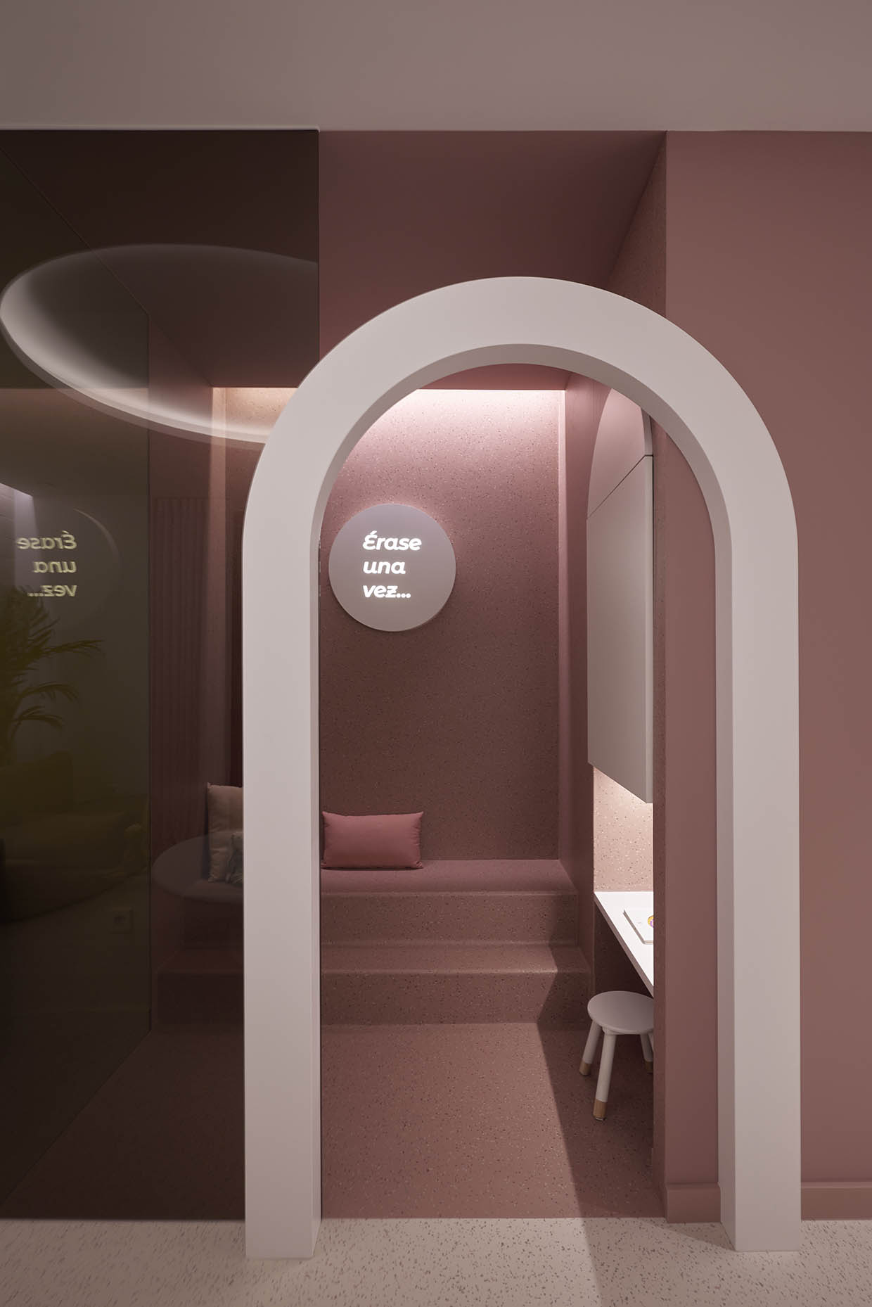 Esta es una zona infantil diseñada con un estilo moderno y acogedor. Se caracteriza por tonos rosados suaves y una iluminación cálida, lo que genera un ambiente tranquilo y acogedor para los niños. El espacio cuenta con un arco blanco que enmarca la entrada, dando una sensación de profundidad y delimitando el área infantil. Dentro, hay un banco empotrado con cojines, creando un rincón cómodo para sentarse y relajarse. También hay una pequeña mesa con un taburete, ideal para actividades como dibujar o leer. En la pared, un cartel iluminado con la frase "Érase una vez..." sugiere que este rincón está destinado a fomentar la imaginación y la lectura. La iluminación indirecta y los materiales de acabado mate refuerzan la sensación de calma y calidez. En general, es un espacio pensado para que los niños se sientan cómodos y seguros, con una estética moderna y funcional.