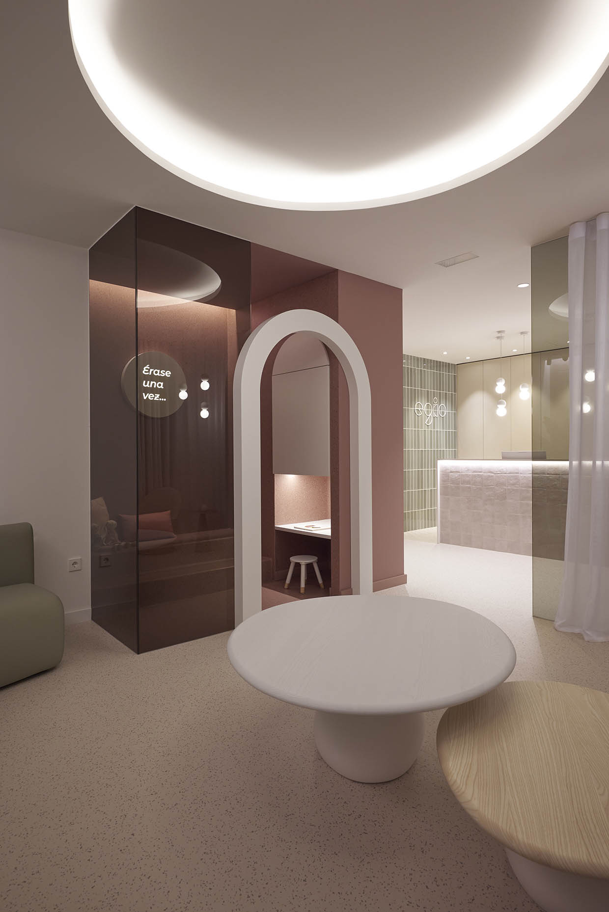 Espacio infantil dentro de la clínica con un diseño acogedor y envolvente. La entrada está enmarcada por un arco blanco que contrasta con las paredes en tonos rosados. En el interior, un banco empotrado con cojines y una pequeña mesa con taburete crean un ambiente ideal para los niños. La iluminación indirecta refuerza la sensación de calidez, mientras que un letrero iluminado con la frase ‘Érase una vez…’ sugiere un espacio lúdico y narrativo, evocando la imaginación y la tranquilidad.