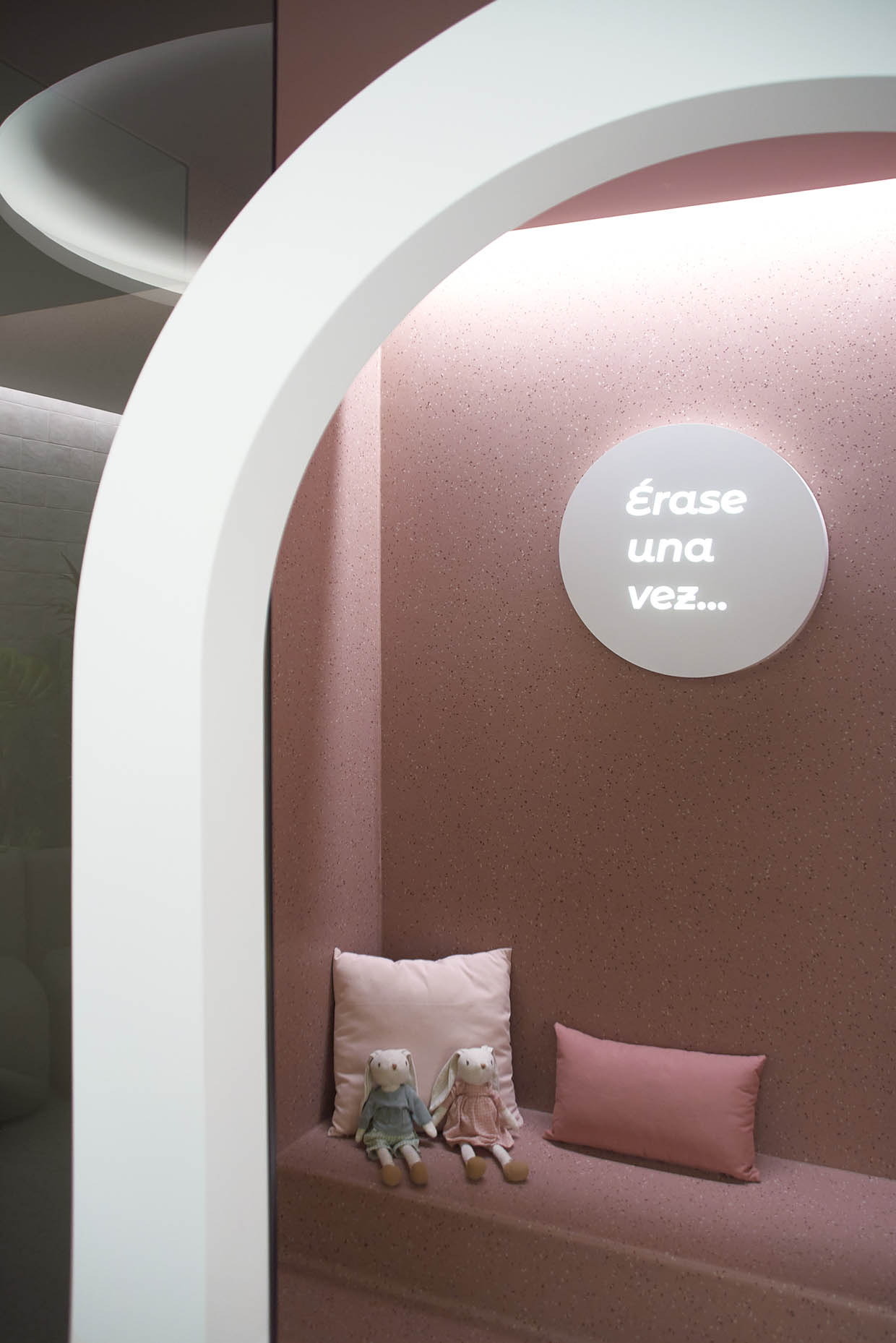 Espacio infantil dentro de la clínica con un diseño acogedor y envolvente. La entrada está enmarcada por un arco blanco que contrasta con las paredes en tonos rosados. En el interior, un banco empotrado con cojines y una pequeña mesa con taburete crean un ambiente ideal para los niños. La iluminación indirecta refuerza la sensación de calidez, mientras que un letrero iluminado con la frase ‘Érase una vez…’ sugiere un espacio lúdico y narrativo, evocando la imaginación y la tranquilidad.