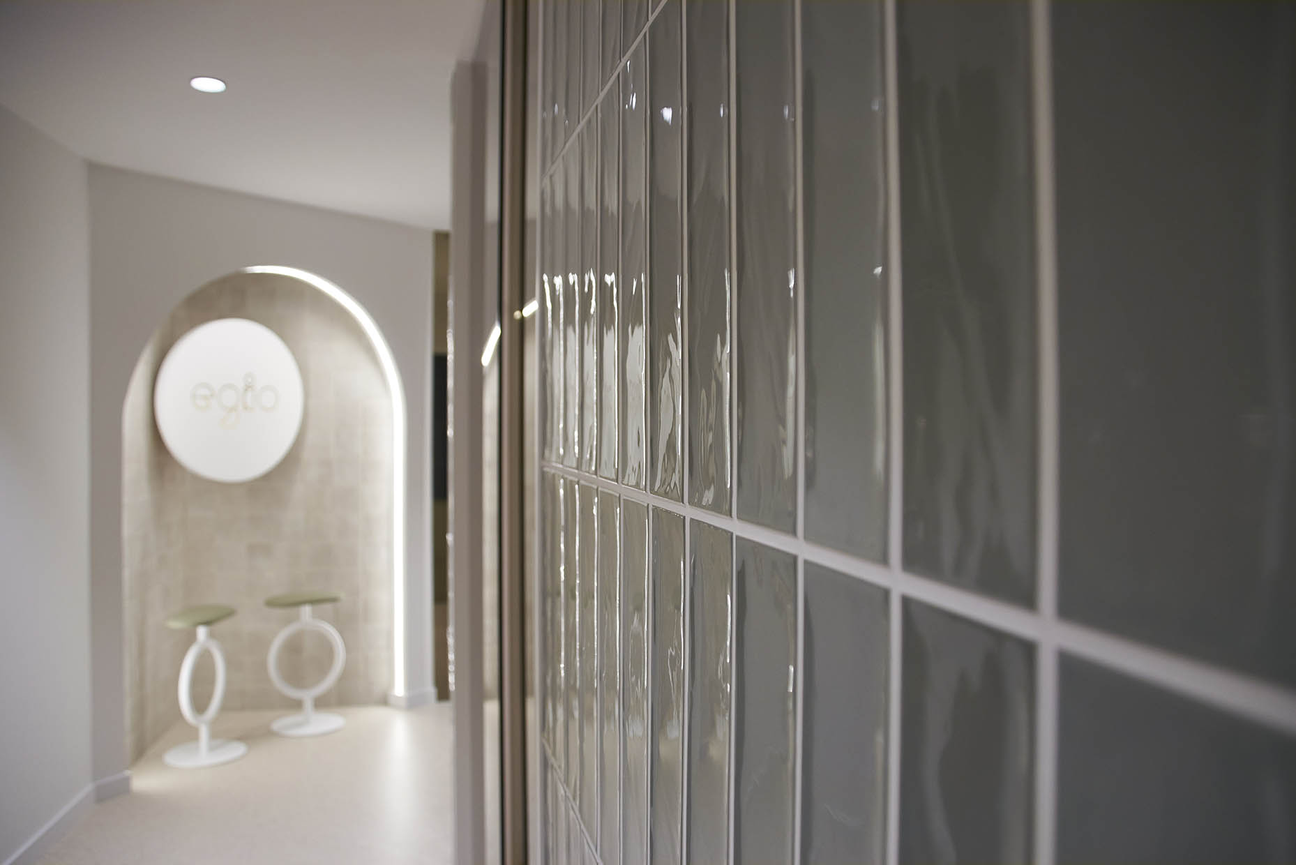 Vista de un pasillo en la clínica con un diseño moderno y minimalista. La pared curva blanca guía el recorrido, mientras que las superficies de vidrio permiten vislumbrar un gabinete odontológico con una silla de tratamiento y un llamativo aro de luz en el techo. Al fondo, un rincón decorativo con un arco iluminado enmarca el logo ‘Egio’, acompañado de dos elegantes taburetes circulares. La iluminación indirecta y los tonos neutros refuerzan la sensación de calma y sofisticación en el espacio.
