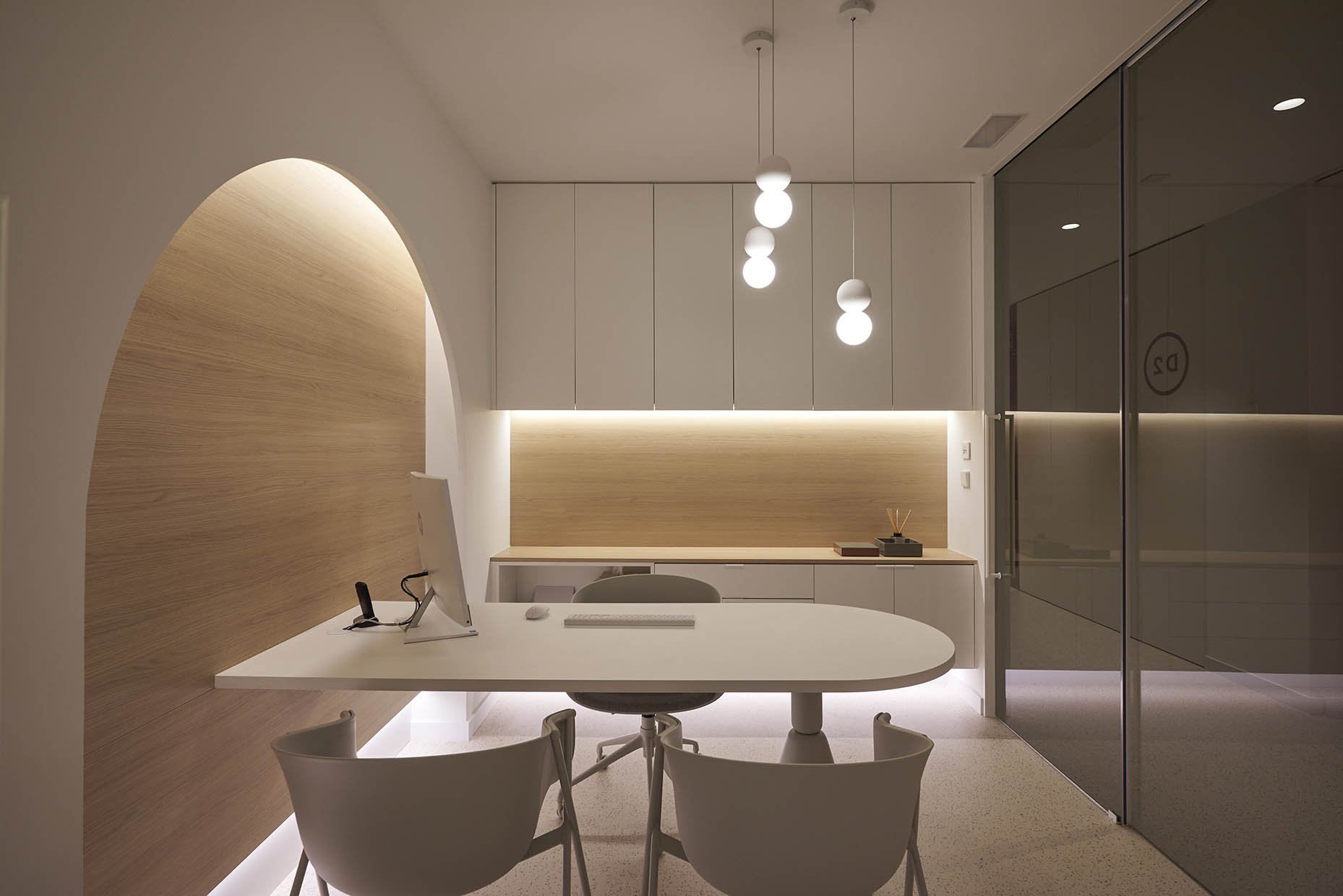 Despacho de consulta en clínica con un diseño elegante y minimalista. El espacio combina tonos blancos y madera clara, con iluminación indirecta para un ambiente cálido y acogedor. Una mesa blanca con sillas modernas crea un área de trabajo funcional, mientras que los armarios superiores ofrecen almacenamiento discreto. Un arco decorativo en la pared añade un toque de diseño distintivo, y las lámparas colgantes aportan iluminación puntual y sofisticación
