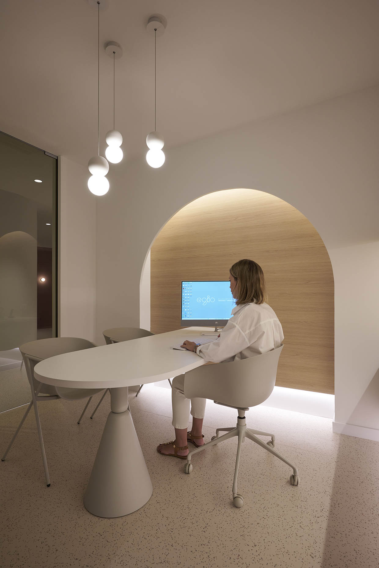 Despacho de consulta en clínica con un diseño elegante y minimalista. El espacio combina tonos blancos y madera clara, con iluminación indirecta para un ambiente cálido y acogedor. Una mesa blanca con sillas modernas crea un área de trabajo funcional, mientras que los armarios superiores ofrecen almacenamiento discreto. Un arco decorativo en la pared añade un toque de diseño distintivo, y las lámparas colgantes aportan iluminación puntual y sofisticación