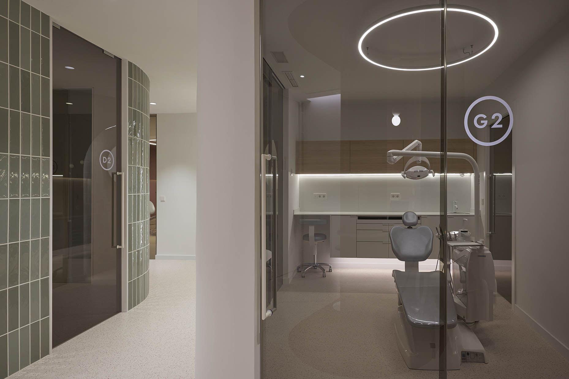 Gabinte dental moderno con un diseño minimalista, donde se destaca un sillón odontológico en tonos grises, rodeado de mobiliario funcional con acabados en madera y superficies blancas. La iluminación es sutil y elegante, con un aro de luz en el techo que aporta un ambiente sofisticado. La sala está separada por puertas de vidrio con rotulación, permitiendo una sensación de amplitud y privacidad. En el pasillo contiguo, se observan paredes revestidas con azulejos verdes y una puerta con cristal ahumado, complementando la estética contemporánea de la clínica