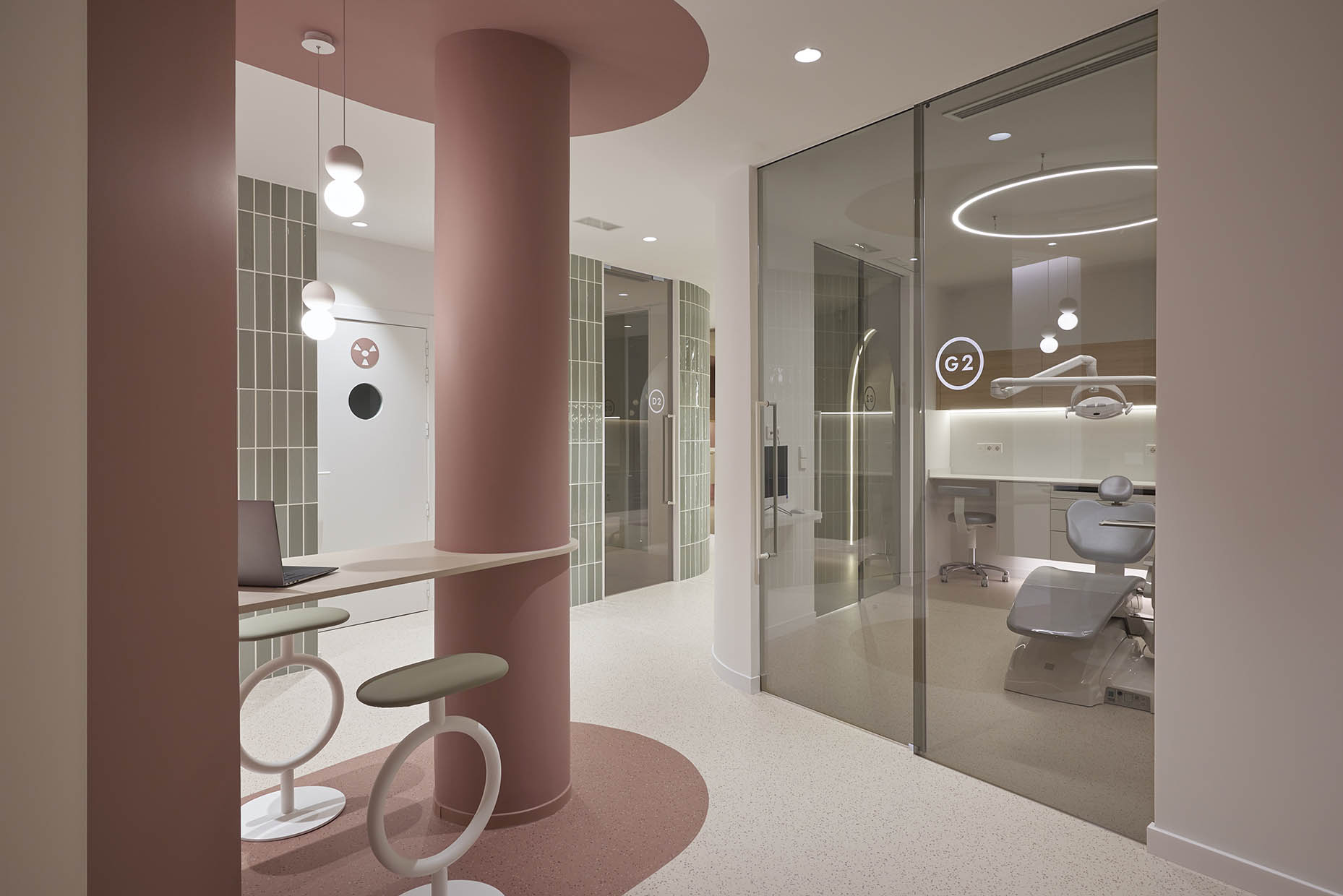 Consultorio dental moderno con un diseño minimalista, donde se destaca un sillón odontológico en tonos grises, rodeado de mobiliario funcional con acabados en madera y superficies blancas. La iluminación es sutil y elegante, con un aro de luz en el techo que aporta un ambiente sofisticado. La sala está separada por puertas de vidrio con rotulación, permitiendo una sensación de amplitud y privacidad. En el pasillo contiguo, se observan paredes revestidas con azulejos verdes y una puerta con cristal ahumado, complementando la estética contemporánea de la clínica.
