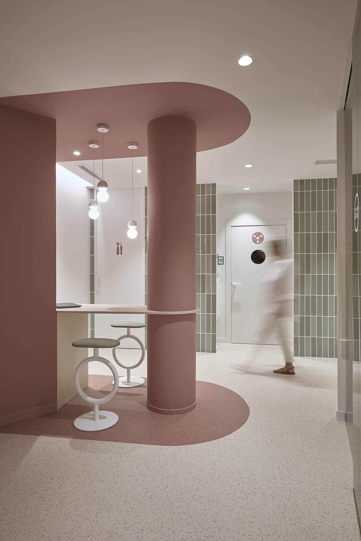 Zona de trabajo dentro de una clínica con un diseño moderno y acogedor. Una mujer con vestimenta blanca trabaja en un ordenador portátil, sentada en un taburete con estructura circular. El espacio está delimitado por una columna cilíndrica y un techo curvo en tono rosa terracota, con iluminación colgante de esferas que aporta calidez. En el fondo, paredes revestidas con azulejos verdes y puertas con señalética minimalista crean un ambiente armonioso y funcional.