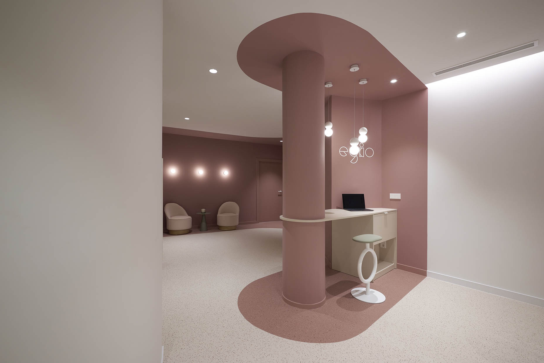 Zona de trabajo dentro de una clínica con un diseño moderno y acogedor. Una mujer con vestimenta blanca trabaja en un ordenador portátil, sentada en un taburete con estructura circular. El espacio está delimitado por una columna cilíndrica y un techo curvo en tono rosa terracota, con iluminación colgante de esferas que aporta calidez. En el fondo, paredes revestidas con azulejos verdes y puertas con señalética minimalista crean un ambiente armonioso y funcional.