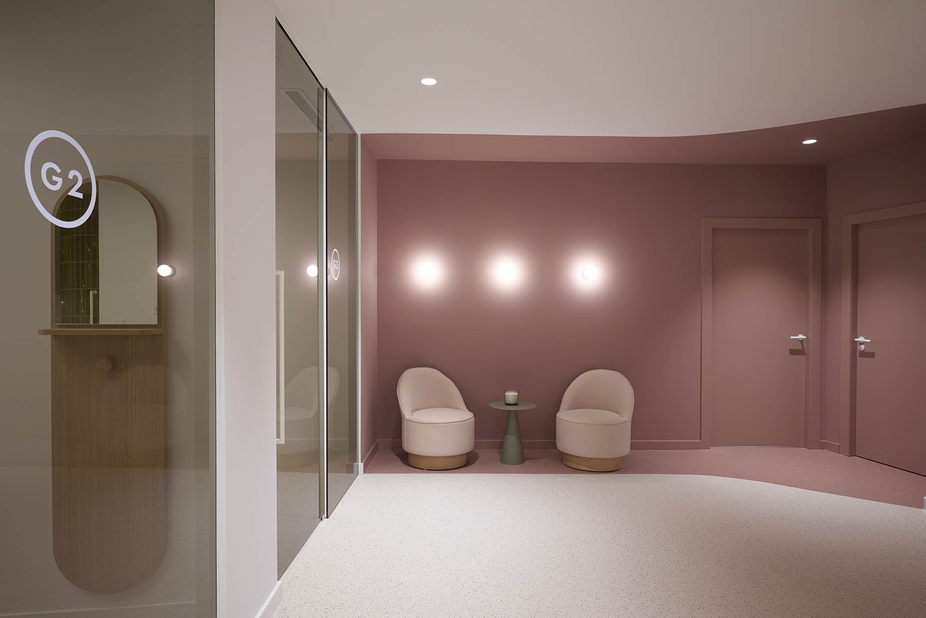 Sala de espera en una clínica con un diseño moderno y minimalista. Predominan tonos rosa empolvado y beige, creando un ambiente acogedor y elegante. Dos sillones curvos tapizados en tono claro se sitúan junto a una pequeña mesa verde con una vela decorativa. La iluminación sutil en la pared genera un efecto relajante.
