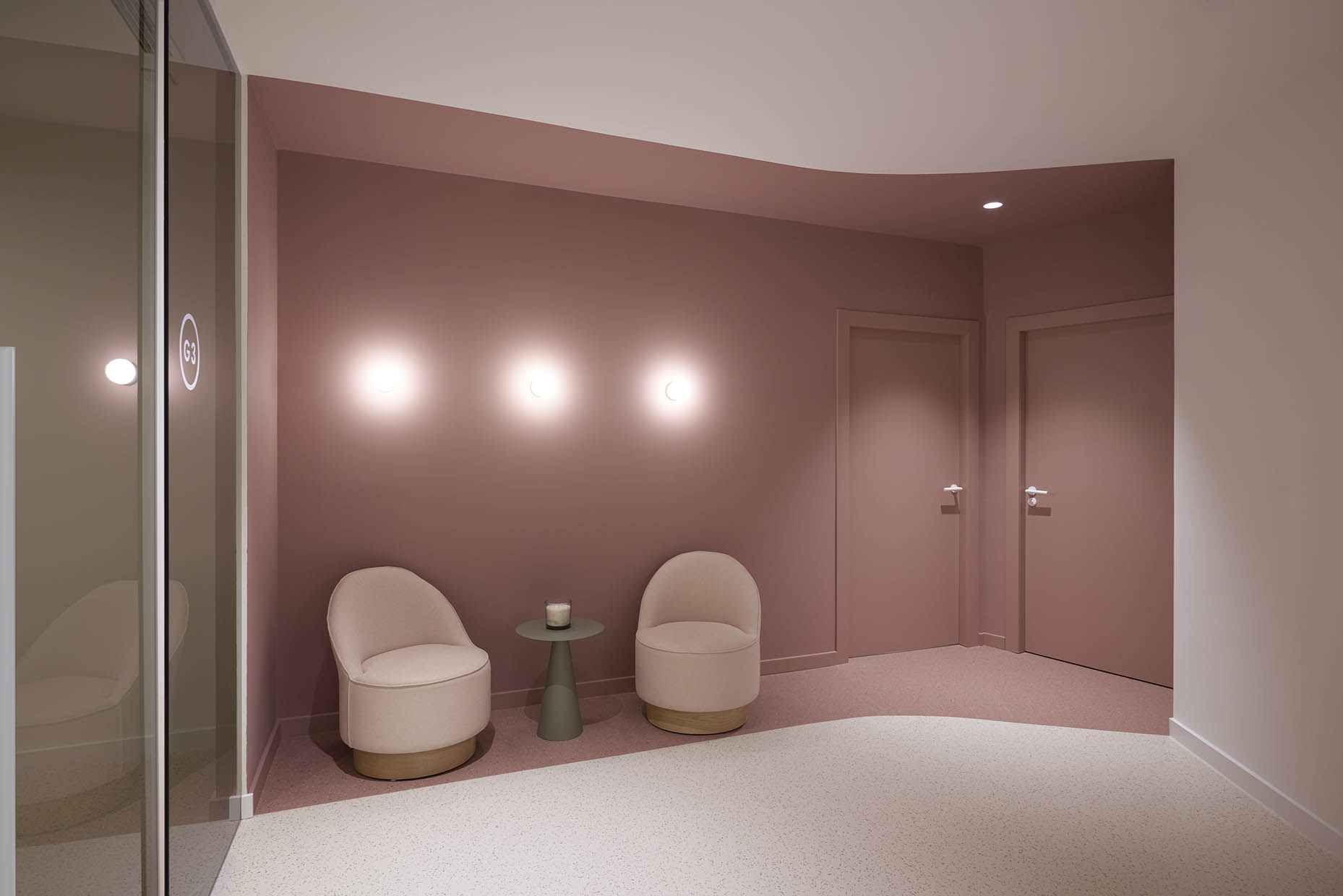 Sala de espera en una clínica con un diseño moderno y minimalista. Predominan tonos rosa empolvado y beige, creando un ambiente acogedor y elegante. Dos sillones curvos tapizados en tono claro se sitúan junto a una pequeña mesa verde con una vela decorativa. La iluminación sutil en la pared genera un efecto relajante.