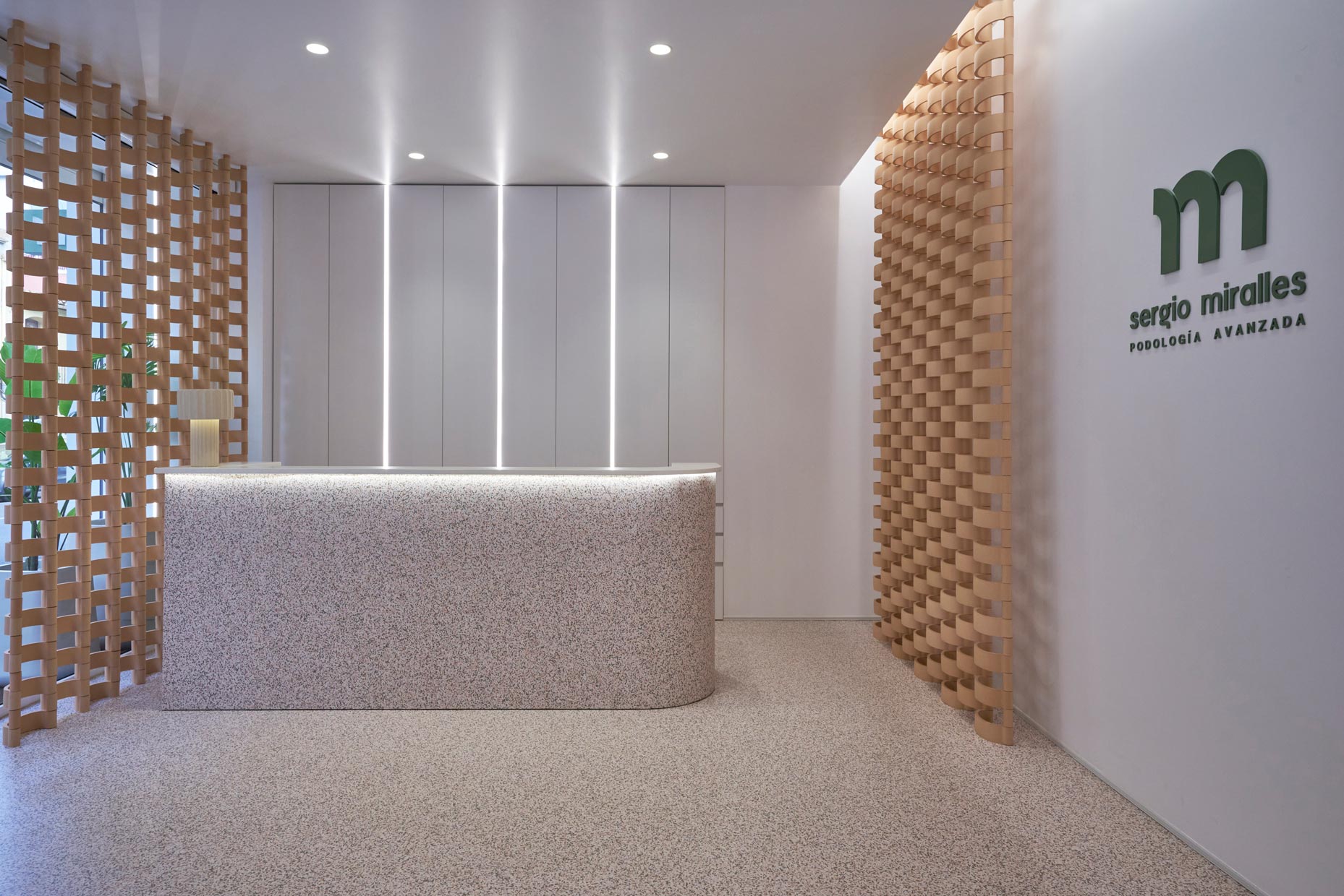 Recepción de clínica con diseño moderno y minimalista. Mostrador curvo revestido de vinilo moteado con iluminación integrada, paredes blancas con líneas de luz verticales y celosías de barro a los lados.
