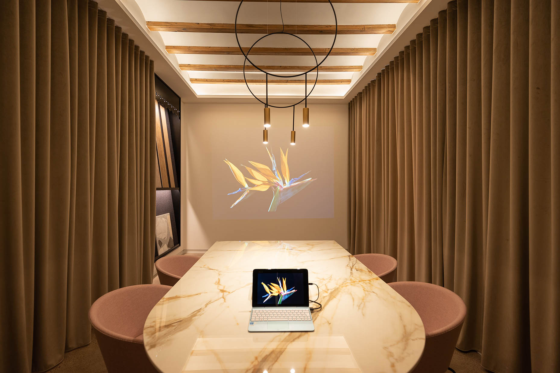Espacio de reunión con una elegante mesa de mármol en tonos beige y vetas doradas, rodeada de sillas tapizadas en rosa empolvado. La iluminación moderna con lámparas colgantes de diseño minimalista y un fondo de cortinas en tono tierra crean un ambiente sofisticado. El techo con vigas de madera y un panel decorativo en la pared con diferentes texturas y materiales aportan calidez y dinamismo al diseño.