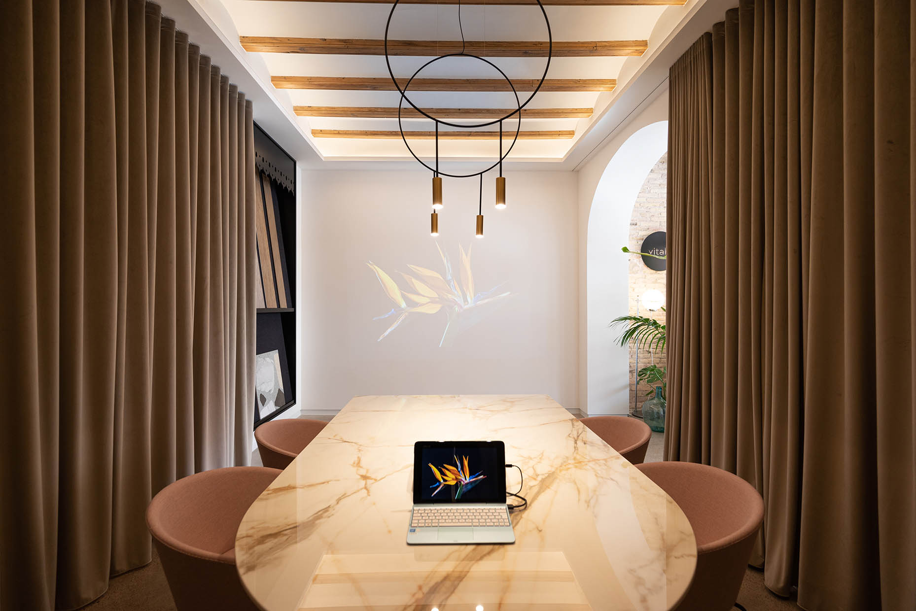 Espacio de reunión con una elegante mesa de mármol en tonos beige y vetas doradas, rodeada de sillas tapizadas en rosa empolvado. La iluminación moderna con lámparas colgantes de diseño minimalista y un fondo de cortinas en tono tierra crean un ambiente sofisticado. El techo con vigas de madera y un panel decorativo en la pared con diferentes texturas y materiales aportan calidez y dinamismo al diseño.