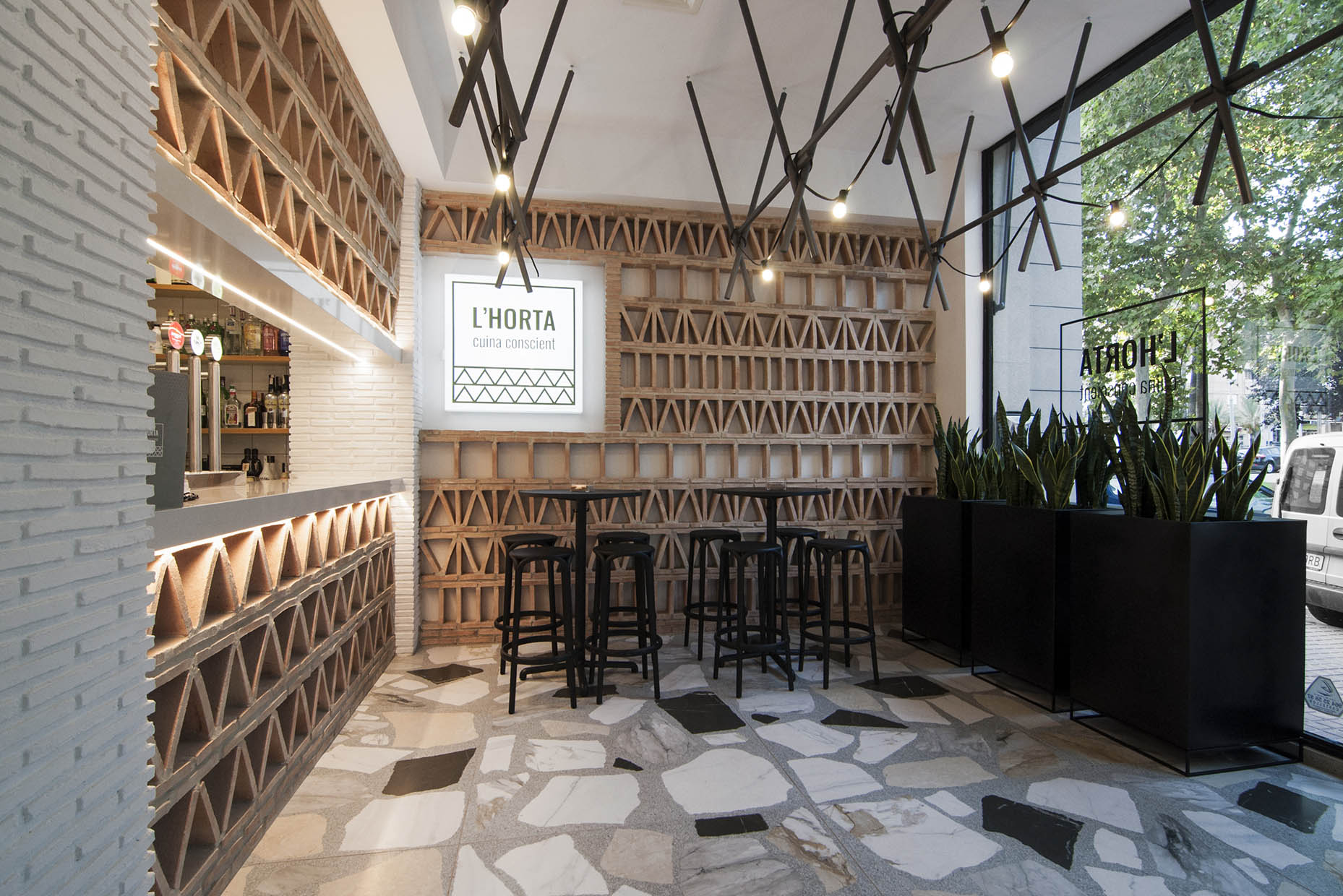 Diseño zona entrada restaurante realizada con ladrillos macizos con colocación según patrones geométricos. Suelo porcelánico de formato 120x120 simulando terrazo. Mobiliario e iluminación suspendida en tonos oscuros. Diseño moderno y elegante.