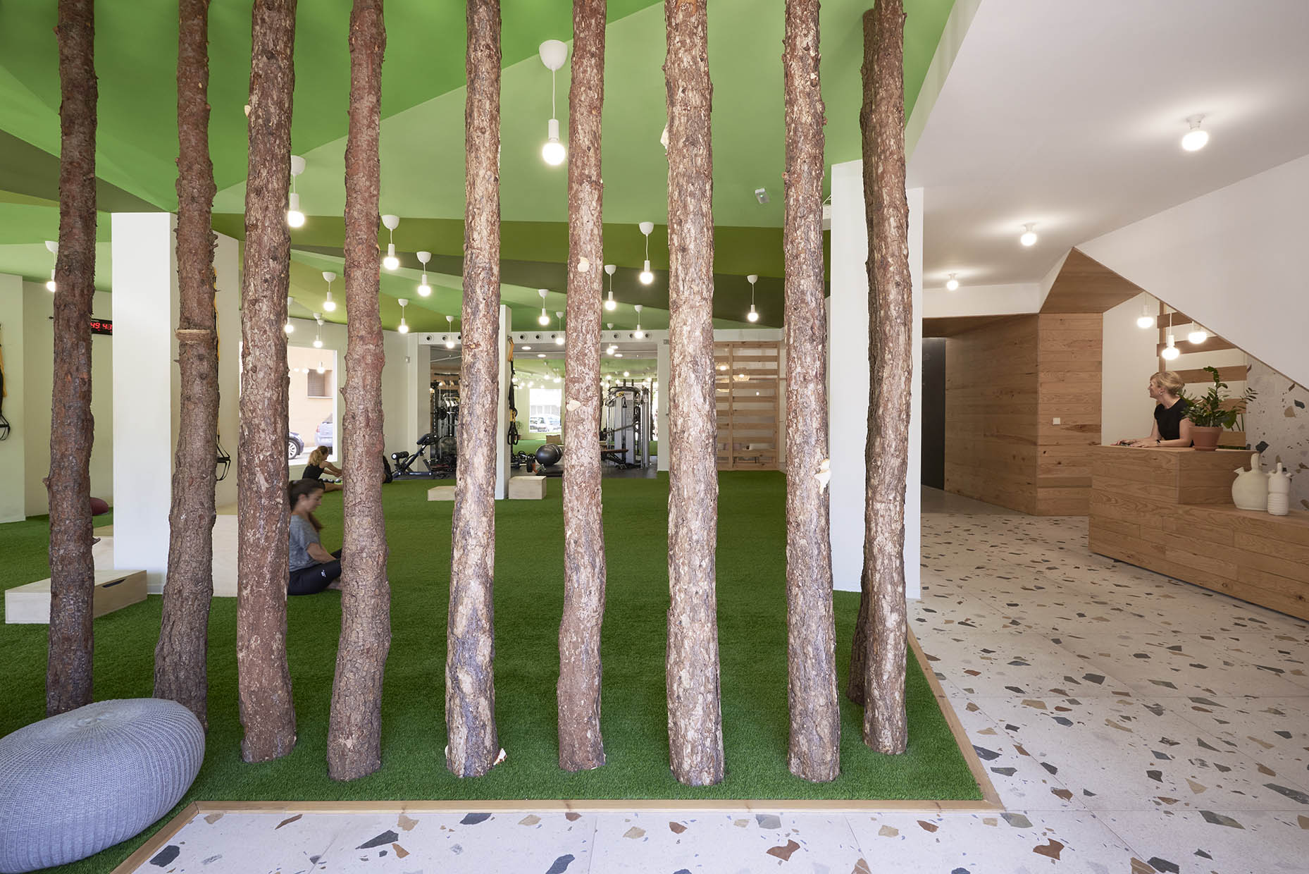 Entrada de un centro de entrenamiento con un diseño inspirado en la naturaleza. El espacio combina troncos de árboles, césped artificial y un techo verde con suelo de terrazo, creando una estética moderna y acogedora.