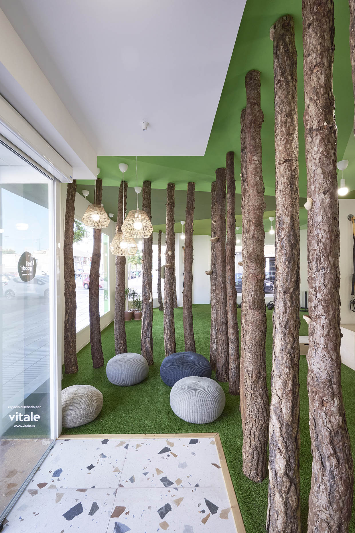 Zona de espera en un centro de entrenamiento con un diseño biofílico. El espacio cuenta con troncos de árboles integrados en la arquitectura, césped artificial en el suelo y un techo verde que refuerza la conexión con la naturaleza. Varias lámparas colgantes de mimbre aportan calidez, mientras que pufs tejidos en tonos neutros invitan a la relajación. Grandes ventanales permiten la entrada de luz natural, creando un ambiente acogedor y equilibrado