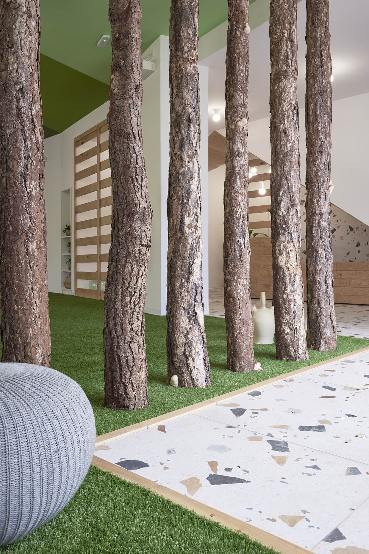 Entrada de un centro de entrenamiento con un diseño inspirado en la naturaleza. El espacio combina troncos de árboles, césped artificial y un techo verde con suelo de terrazo, creando una estética moderna y acogedora.