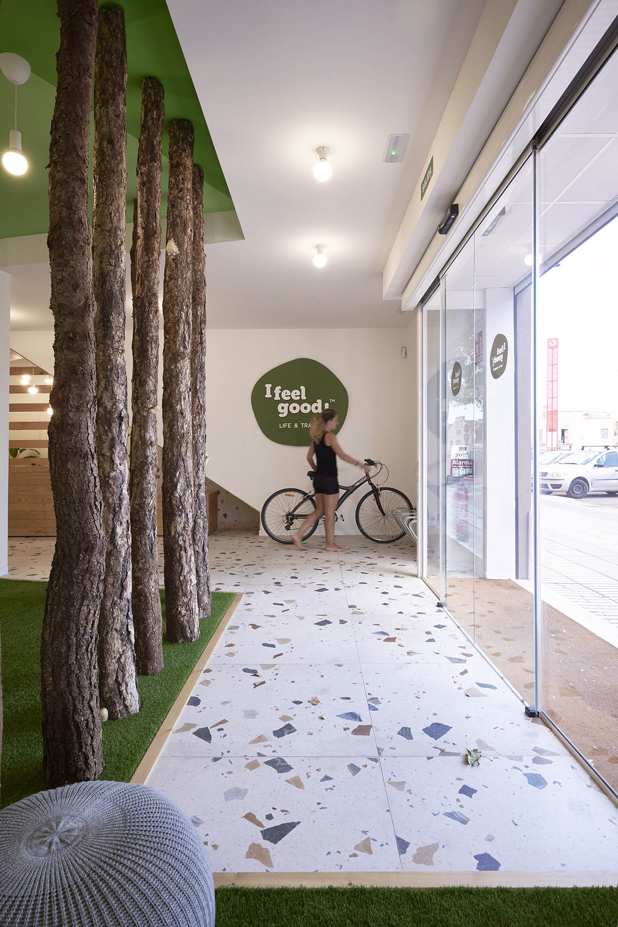 Entrada de un centro de entrenamiento con un diseño inspirado en la naturaleza. El espacio combina troncos de árboles, césped artificial y un techo verde con suelo de terrazo, creando una estética moderna y acogedora. Una mujer descalza camina junto a su bicicleta hacia la salida de cristal, que permite la entrada de luz natural. En la pared, un letrero verde con la frase 'I feel good. LIFE & TRAINING' refuerza el concepto de bienestar del centro