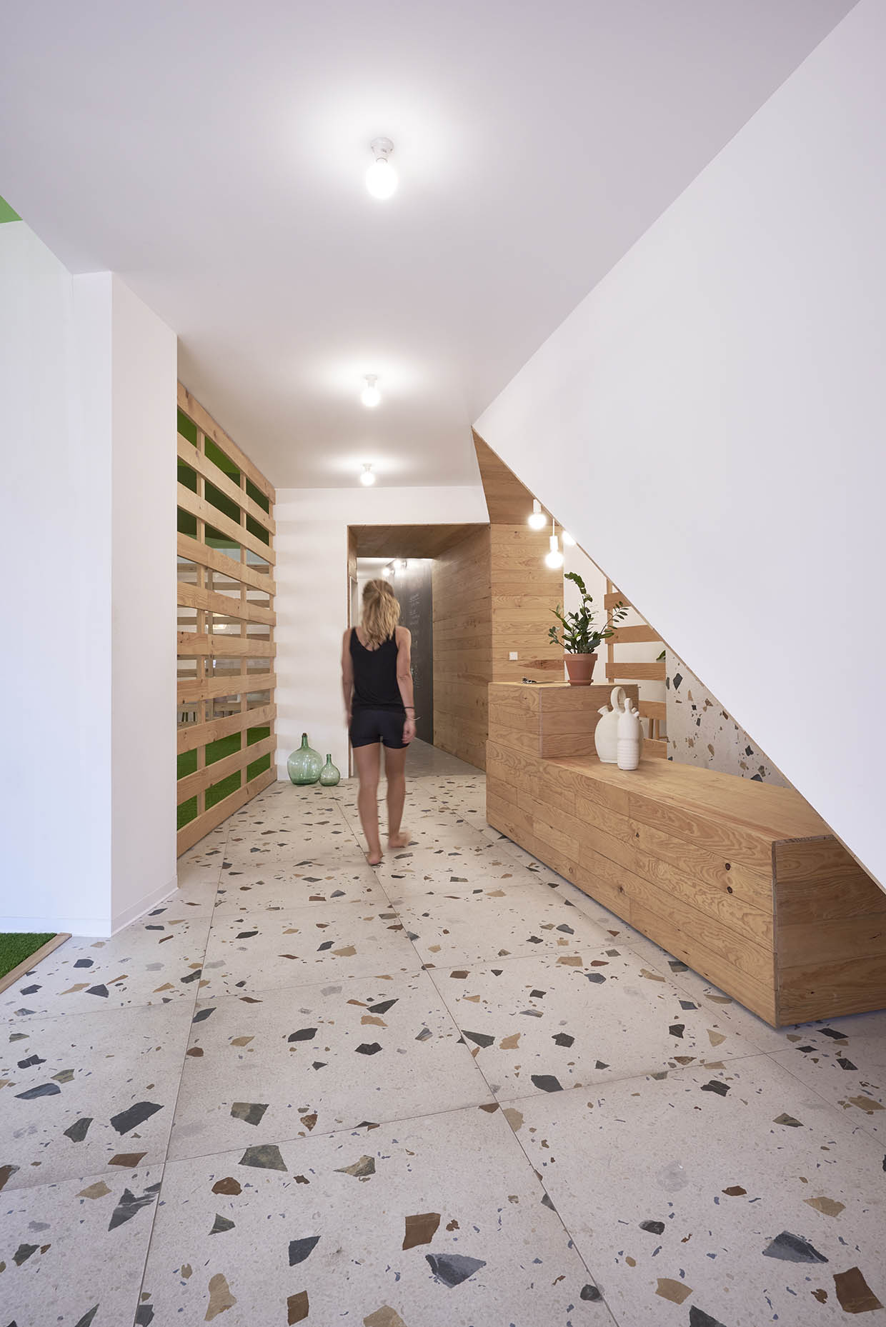 nterior de un centro de entrenamiento con un diseño cálido y natural. Una mujer descalza y vestida de negro camina por un pasillo con suelo de terrazo, paredes blancas y detalles en madera. A la derecha, un mostrador de recepción de madera con decoración minimalista y luces colgantes. A la izquierda, una estructura de listones de madera con fondo verde aporta un toque orgánico al espacio. El ambiente es luminoso y acogedor, transmitiendo una sensación de bienestar