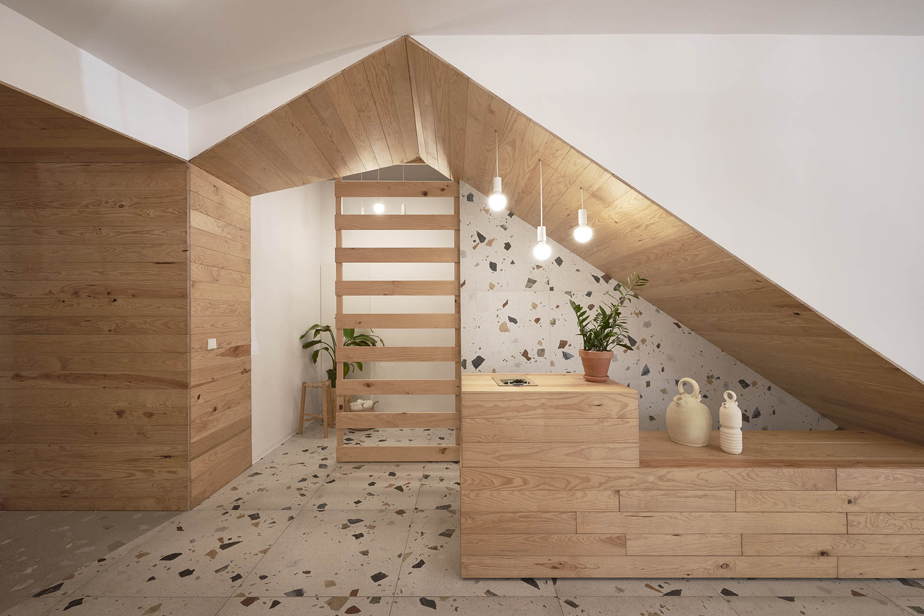 Recepción de un centro de entrenamiento con un diseño acogedor y natural. Un mostrador de madera clara se integra armoniosamente bajo una escalera revestida del mismo material. El fondo combina paredes blancas con revestimiento de terrazo, aportando textura y calidez. Tres lámparas colgantes con bombillas expuestas iluminan suavemente el espacio. Sobre el mostrador, hay una planta en una maceta de barro y dos jarrones de cerámica de diseño artesanal. A la izquierda, un panel de listones de madera separa visualmente otra zona, acompañado de una pequeña planta en un taburete. El ambiente es minimalista y elegante, con un estilo natural y relajante.