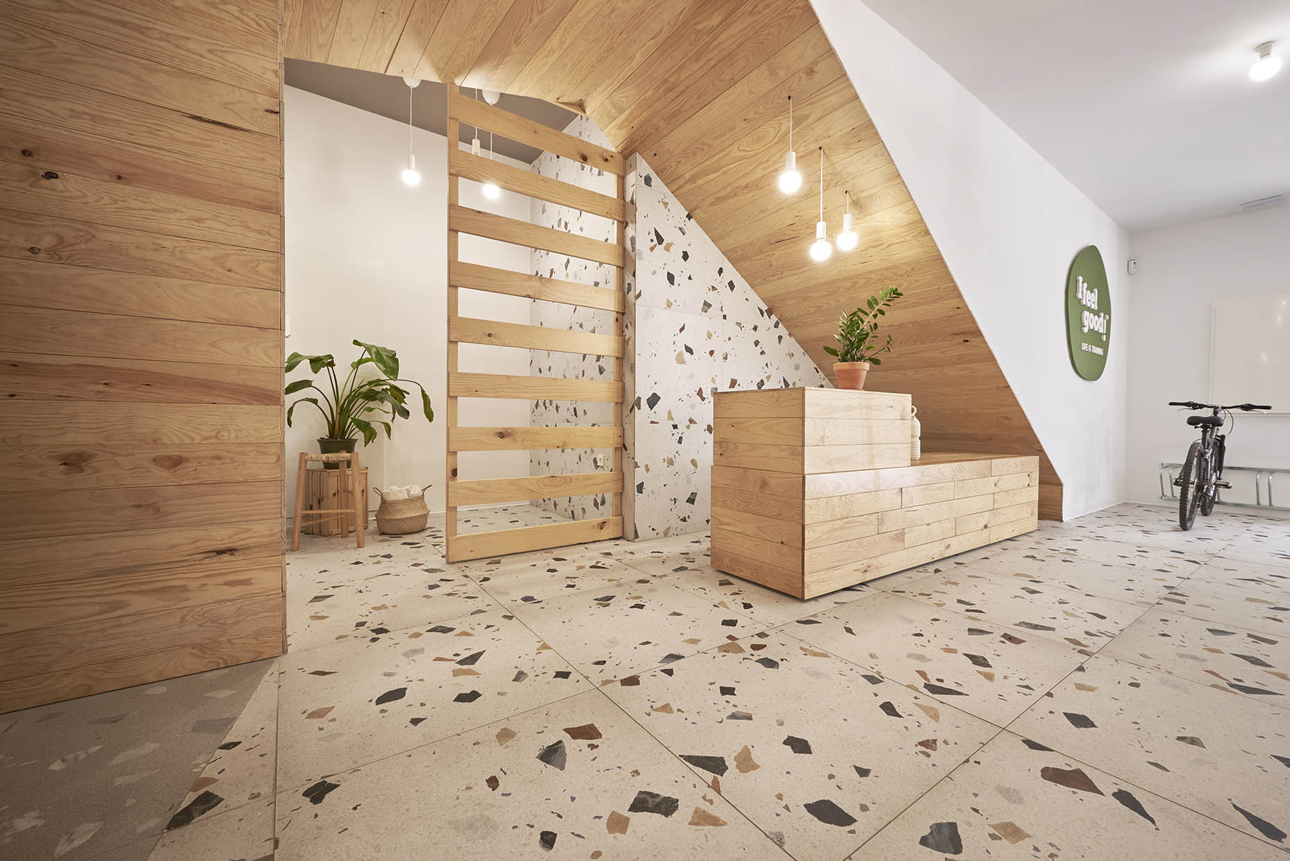 Recepción de un centro de entrenamiento con un diseño acogedor y natural. Un mostrador de madera clara se integra armoniosamente bajo una escalera revestida del mismo material. El fondo combina paredes blancas con revestimiento de terrazo, aportando textura y calidez. Tres lámparas colgantes con bombillas expuestas iluminan suavemente el espacio. Sobre el mostrador, hay una planta en una maceta de barro y dos jarrones de cerámica de diseño artesanal. A la izquierda, un panel de listones de madera separa visualmente otra zona, acompañado de una pequeña planta en un taburete. El ambiente es minimalista y elegante, con un estilo natural y relajante.