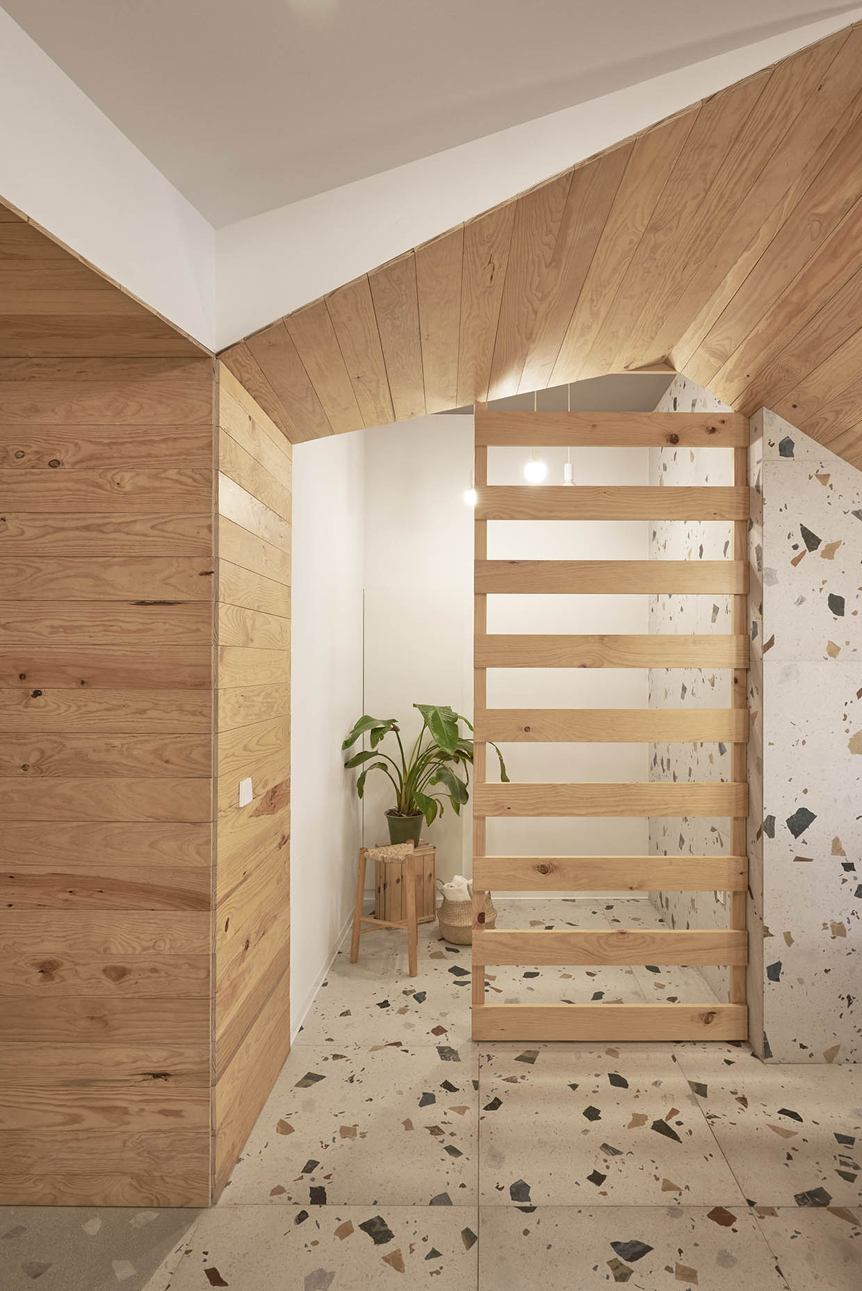 Recepción de un centro de entrenamiento con un diseño acogedor y natural. Un mostrador de madera clara se integra armoniosamente bajo una escalera revestida del mismo material. El fondo combina paredes blancas con revestimiento de terrazo, aportando textura y calidez. Tres lámparas colgantes con bombillas expuestas iluminan suavemente el espacio. Sobre el mostrador, hay una planta en una maceta de barro y dos jarrones de cerámica de diseño artesanal. A la izquierda, un panel de listones de madera separa visualmente otra zona, acompañado de una pequeña planta en un taburete. El ambiente es minimalista y elegante, con un estilo natural y relajante.