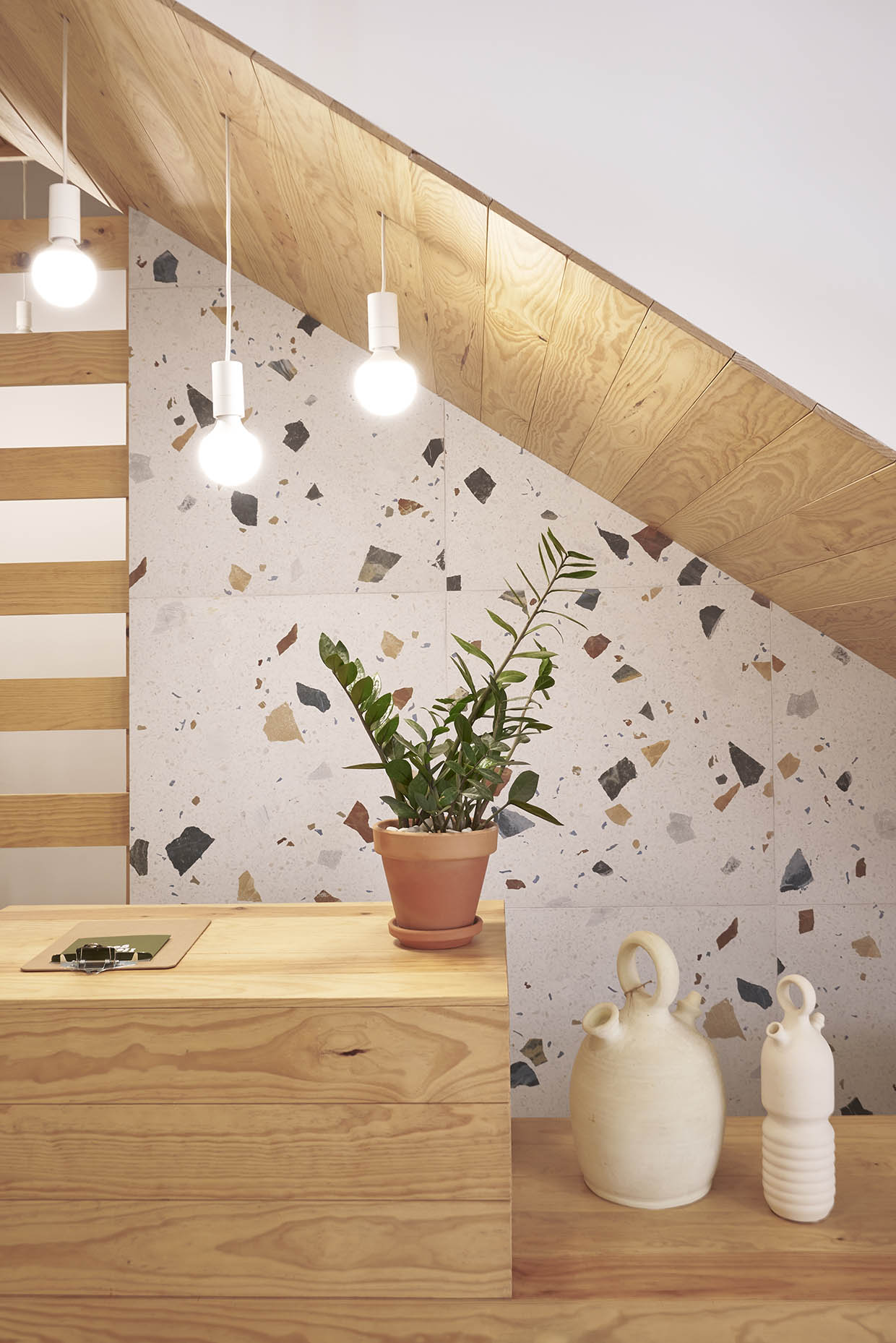 Recepción de un centro de entrenamiento con un diseño acogedor y natural. Un mostrador de madera clara se integra armoniosamente bajo una escalera revestida del mismo material. El fondo combina paredes blancas con revestimiento de terrazo, aportando textura y calidez. Tres lámparas colgantes con bombillas expuestas iluminan suavemente el espacio. Sobre el mostrador, hay una planta en una maceta de barro y dos jarrones de cerámica de diseño artesanal. A la izquierda, un panel de listones de madera separa visualmente otra zona, acompañado de una pequeña planta en un taburete. El ambiente es minimalista y elegante, con un estilo natural y relajante.