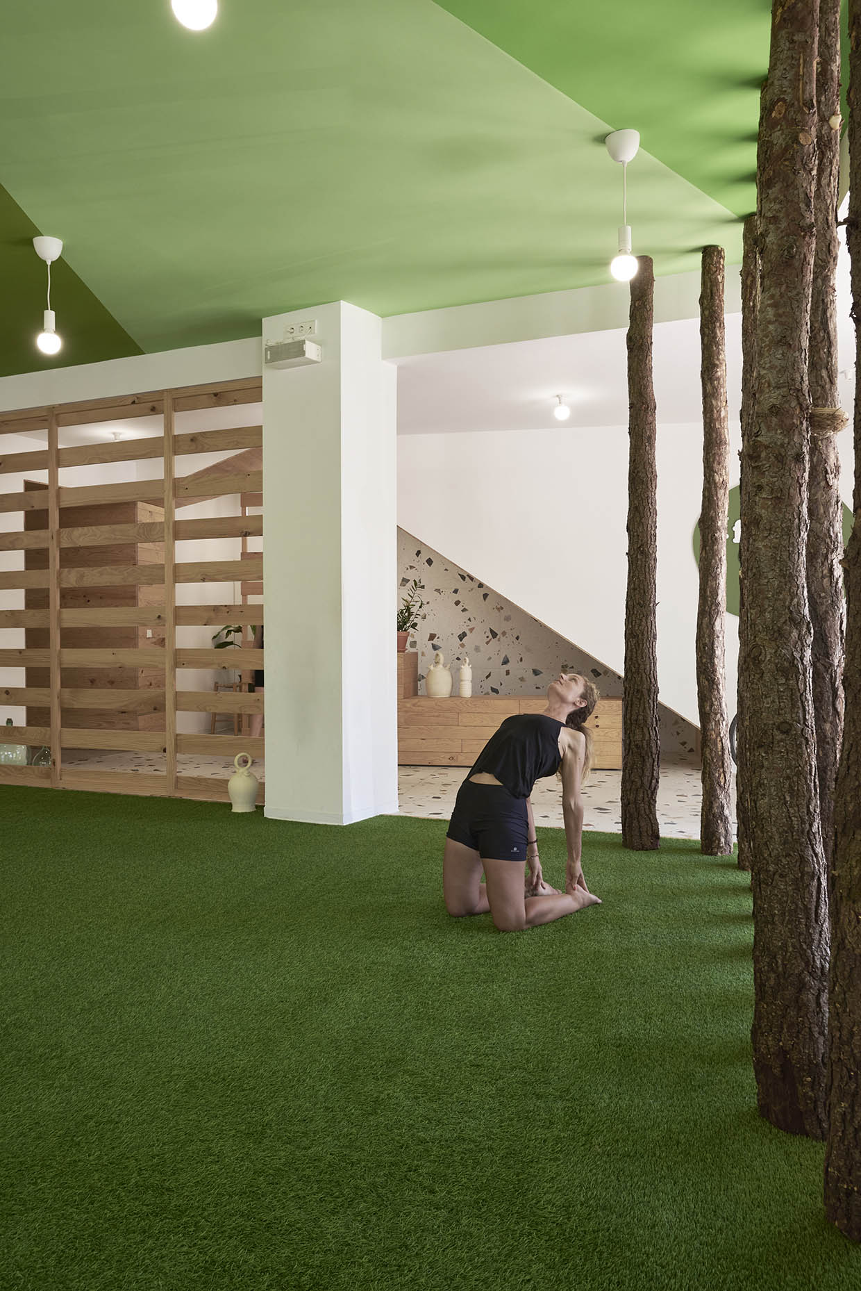 Espacio de entrenamiento con una estética natural y relajante. El suelo está cubierto de césped artificial, integrando elementos que evocan un entorno al aire libre. Varios troncos de árbol se alzan desde el suelo hasta el techo verde, creando una atmósfera forestal dentro del espacio