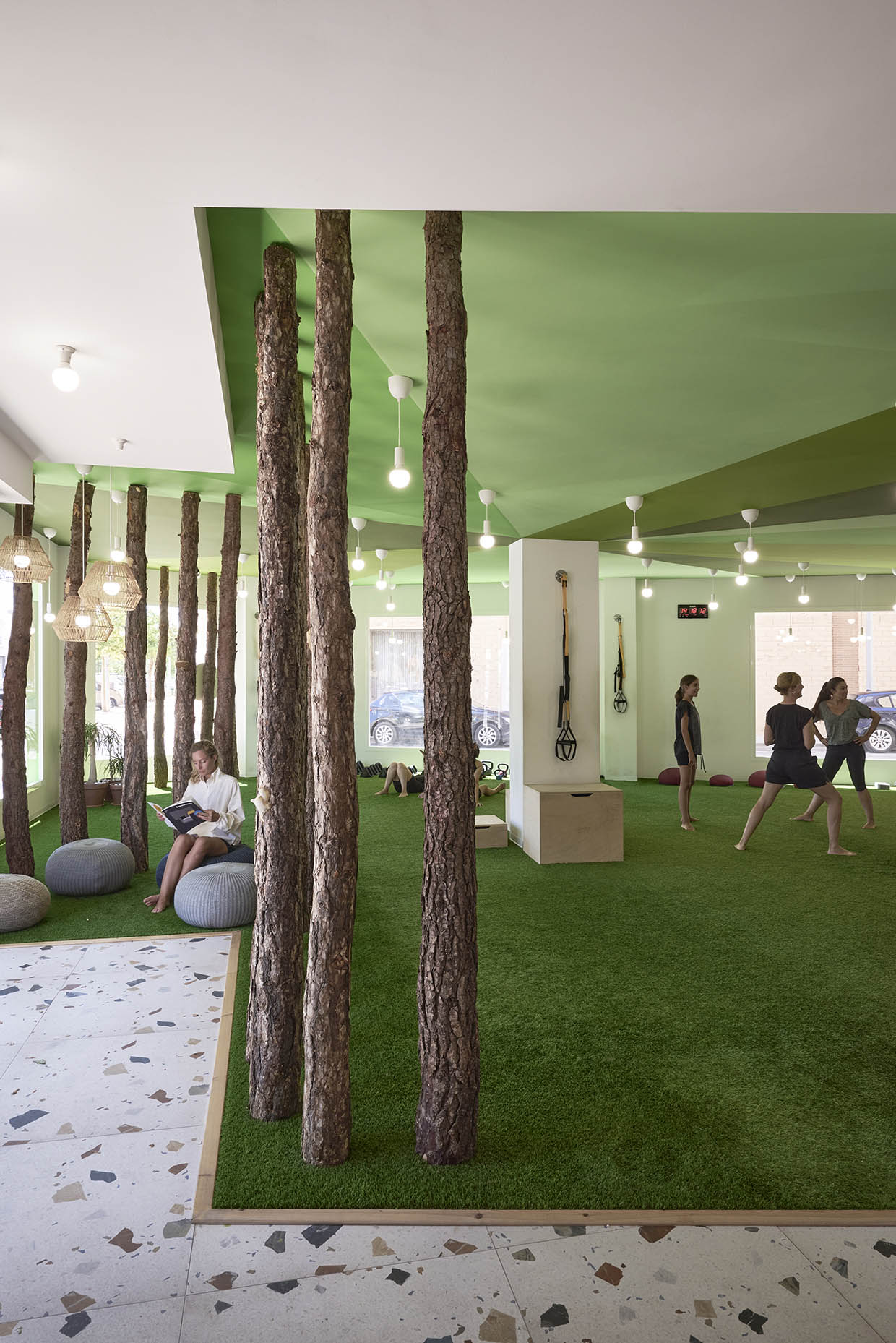 Espacio de entrenamiento con una estética natural y relajante. El suelo está cubierto de césped artificial, integrando elementos que evocan un entorno al aire libre. Varios troncos de árbol se alzan desde el suelo hasta el techo verde, creando una atmósfera forestal dentro del espacio. A la izquierda, una persona está sentada sobre un puf, leyendo tranquilamente. Al fondo, un grupo de mujeres practica ejercicios funcionales y estiramientos descalzas, rodeadas de equipo de entrenamiento como pesas, TRX y cajones de salto. Grandes ventanales permiten la entrada de luz natural, mientras que lámparas colgantes de diseño minimalista iluminan el área. El diseño fomenta una conexión con la naturaleza, combinando bienestar físico y mental