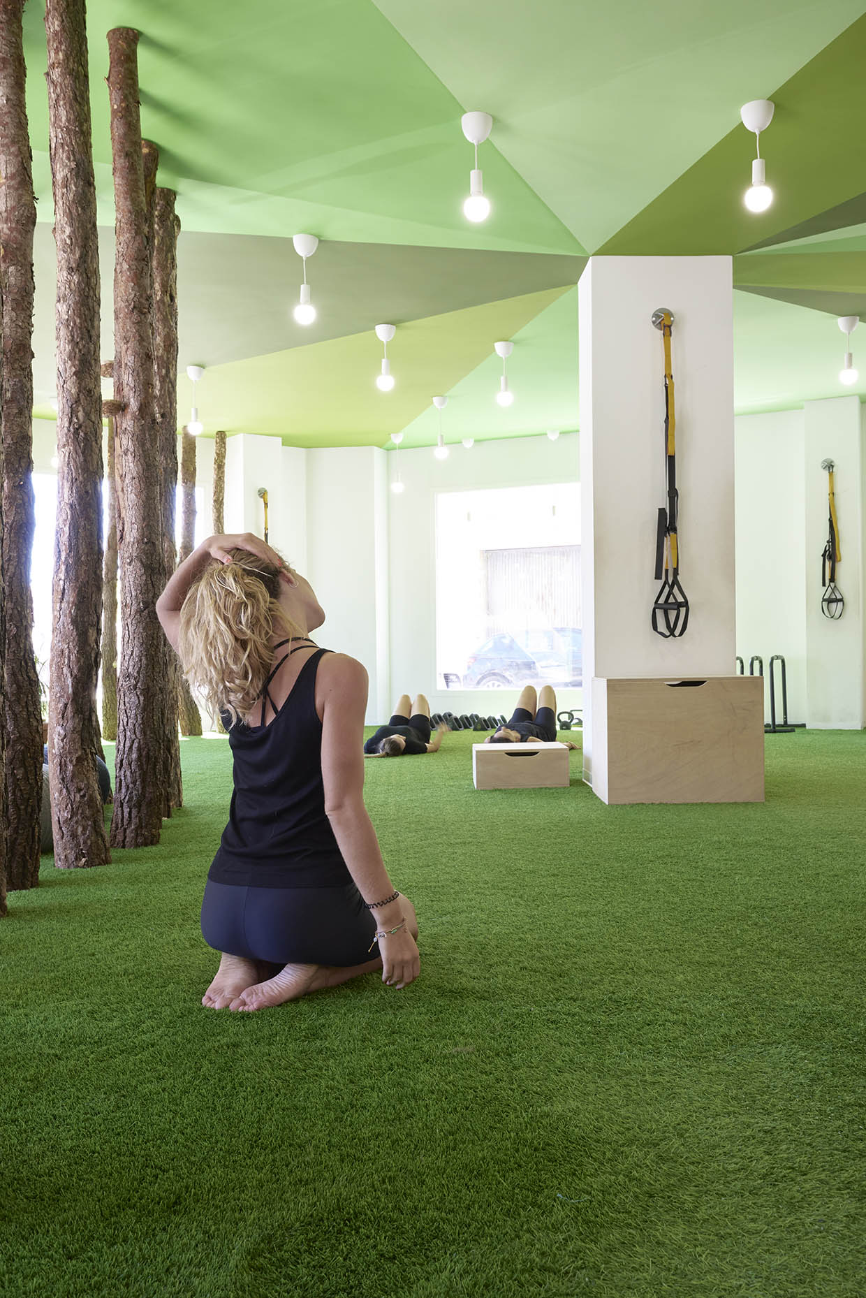 Espacio de entrenamiento con una estética natural y relajante. El suelo está cubierto de césped artificial, integrando elementos que evocan un entorno al aire libre. Varios troncos de árbol se alzan desde el suelo hasta el techo verde, creando una atmósfera forestal dentro del espacio. Grupo de mujeres estirando sobre el suelo de cesped.