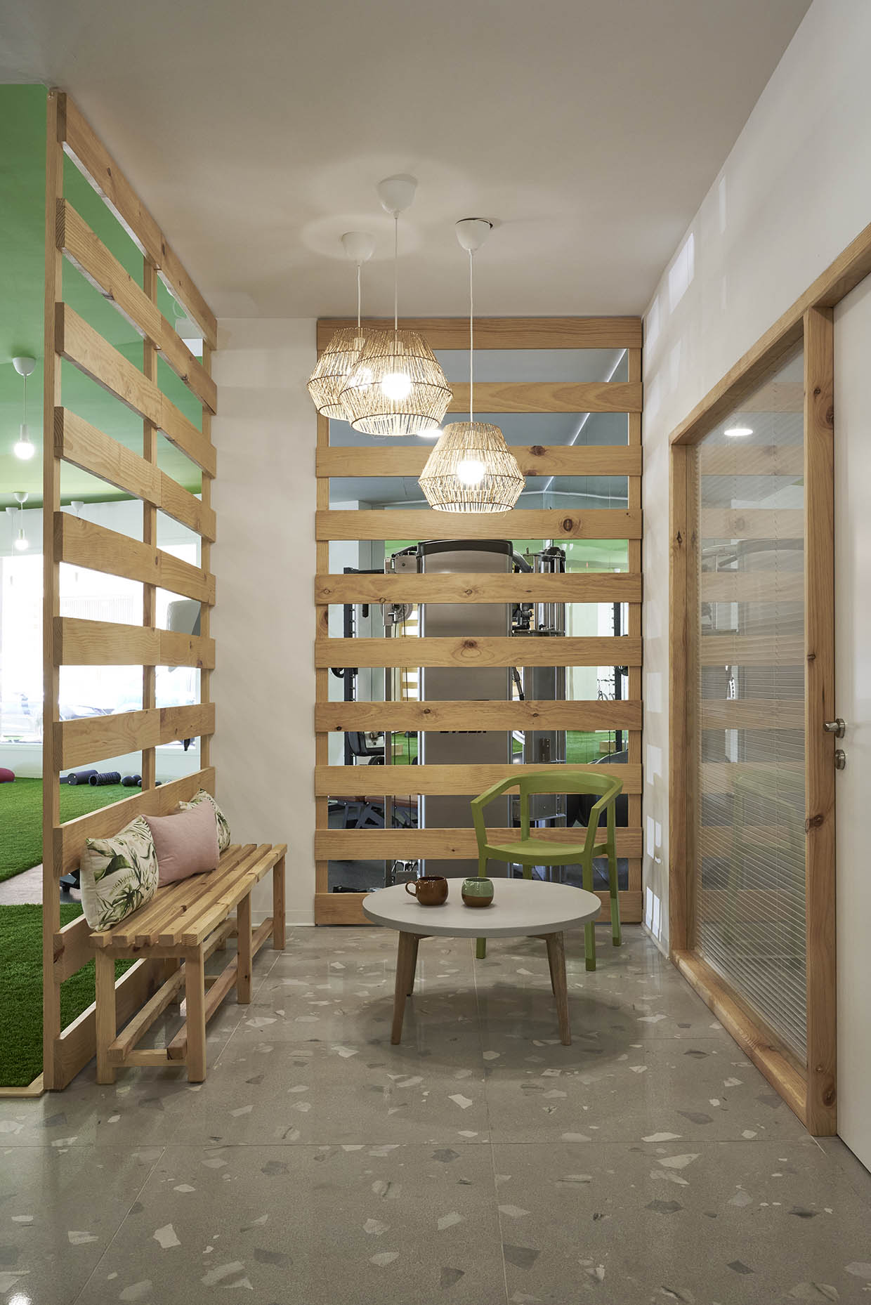 Acogedora zona de espera con un diseño natural y minimalista. El espacio está delimitado por listones de madera que permiten la visibilidad hacia el gimnasio, integrando ambas áreas. Un banco de madera con cojines decorativos en tonos tierra y verdes aporta comodidad. Una pequeña mesa redonda con tazas de cerámica sugiere un ambiente relajante. La silla verde añade un toque de color, complementando la temática natural del diseño. Sobre la mesa, tres lámparas colgantes de mimbre generan una iluminación cálida y acogedora. El suelo de terrazo gris y las paredes blancas equilibran el conjunto, mientras que una puerta de madera con cristal translúcido brinda acceso a otra área del recinto.