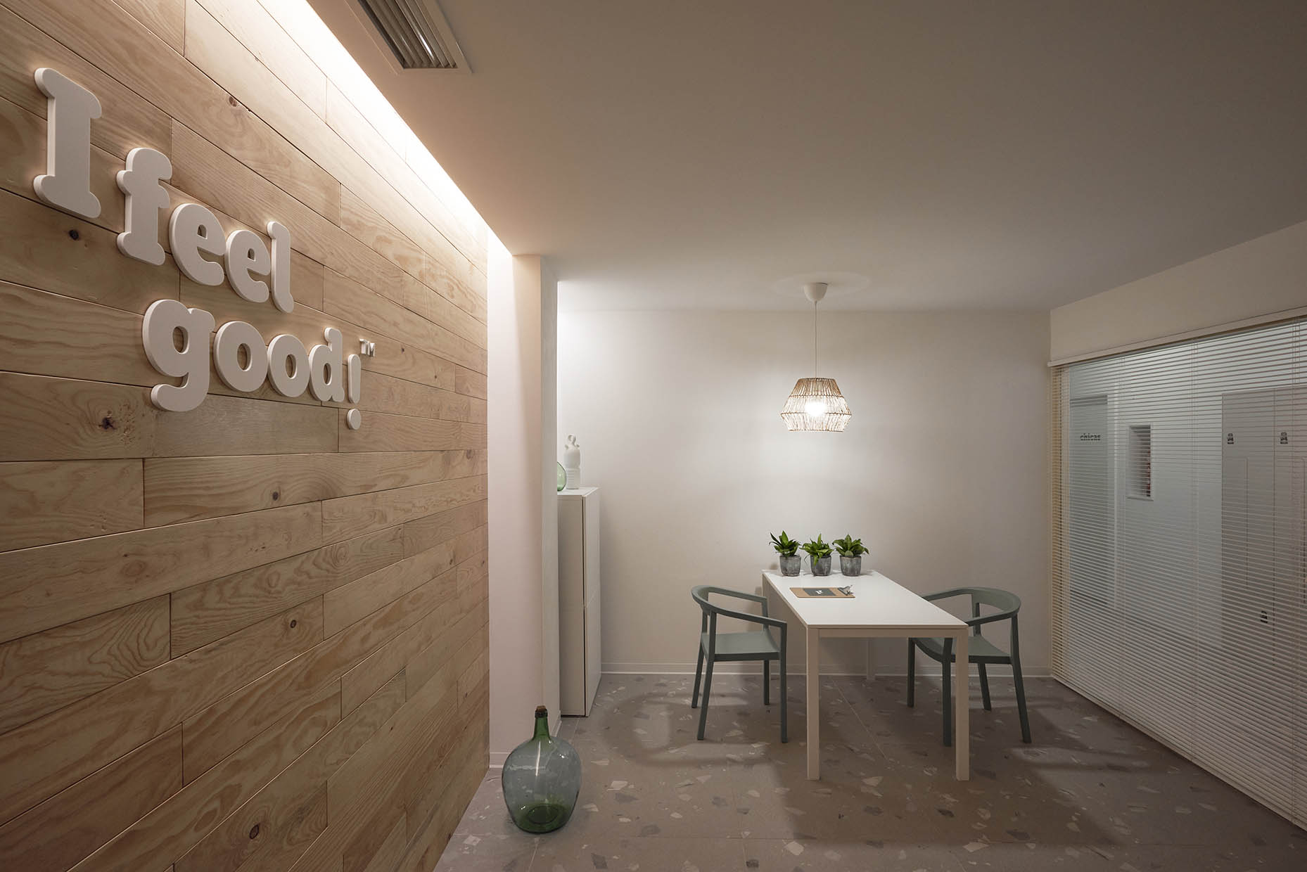 Diseño de gabinete de fisioterapia con un diseño minimalista y acogedor. En la pared de madera clara destaca el mensaje 'I feel good!' en letras en relieve. El espacio cuenta con una mesa blanca acompañada de dos sillas de diseño moderno en tono verde, decorada con pequeñas macetas con plantas. Una lámpara colgante de mimbre proporciona iluminación cálida. Al fondo, una pared de cristal con persianas permite vislumbrar otras áreas del centro, manteniendo privacidad y amplitud visual. El ambiente combina materiales naturales y tonos neutros para transmitir serenidad y bienestar.